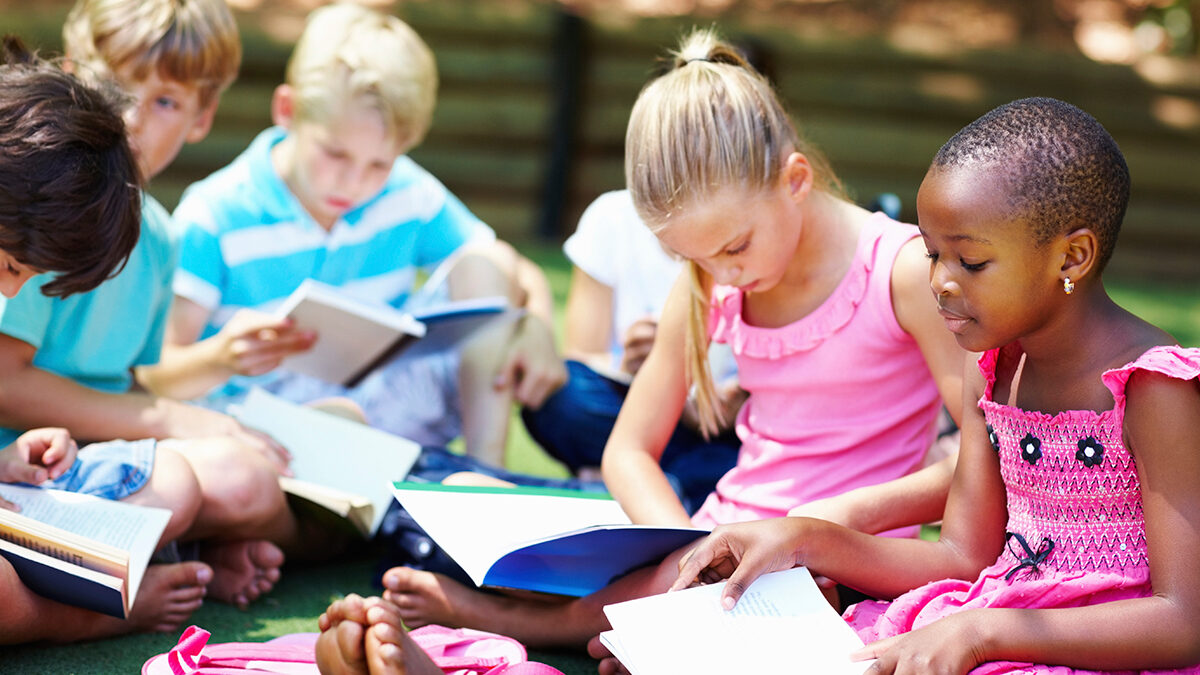 Children sitting outdoors reading books together as part of community support services that encourage learning and connection.