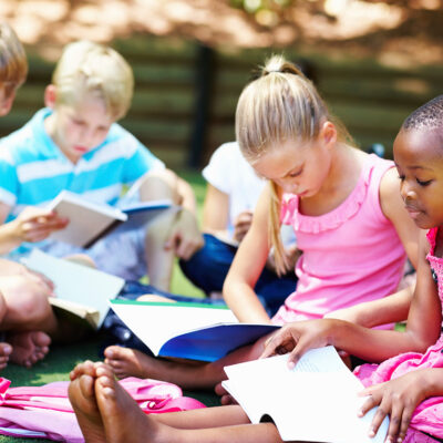 Children sitting outdoors reading books together as part of community support services that encourage learning and connection.