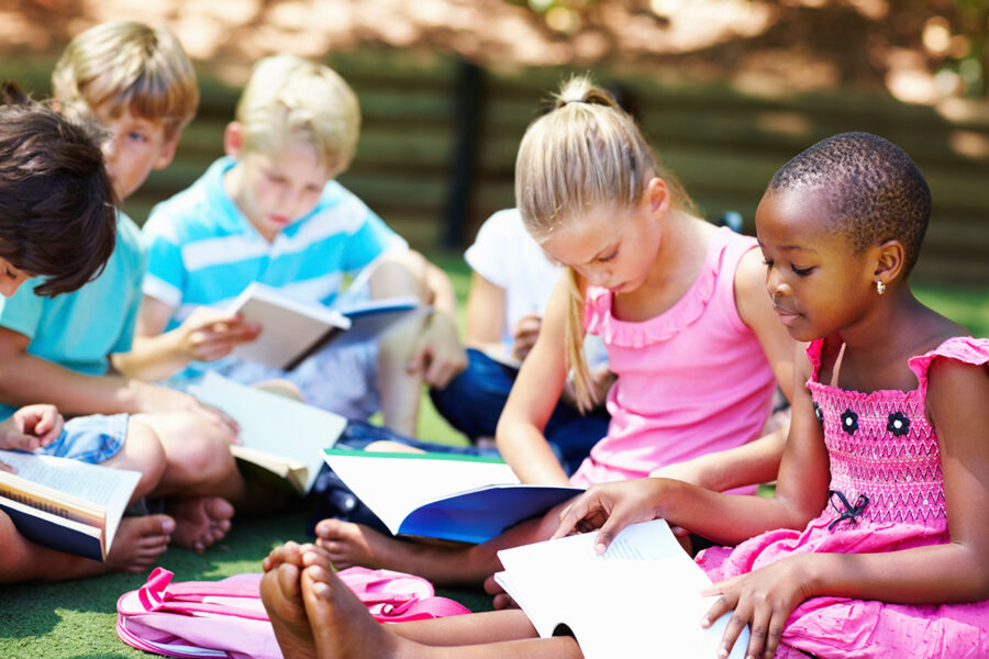 Children sitting outdoors reading books together as part of community support services that encourage learning and connection.