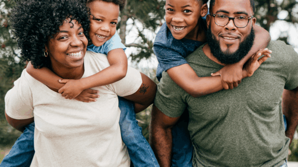 A mother and father smiling carrying a child on each of their backs.