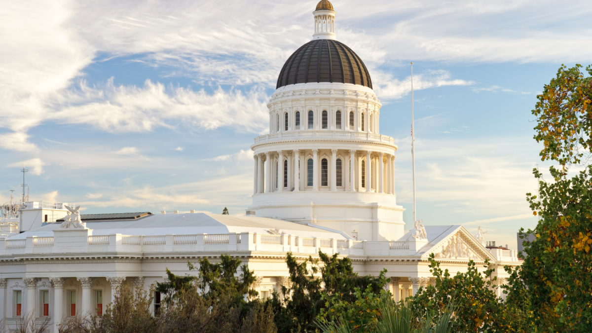 View of the California State Capitol building in Sacramento under a bright sky with wispy clouds, framed by trees and greenery in the foreground.