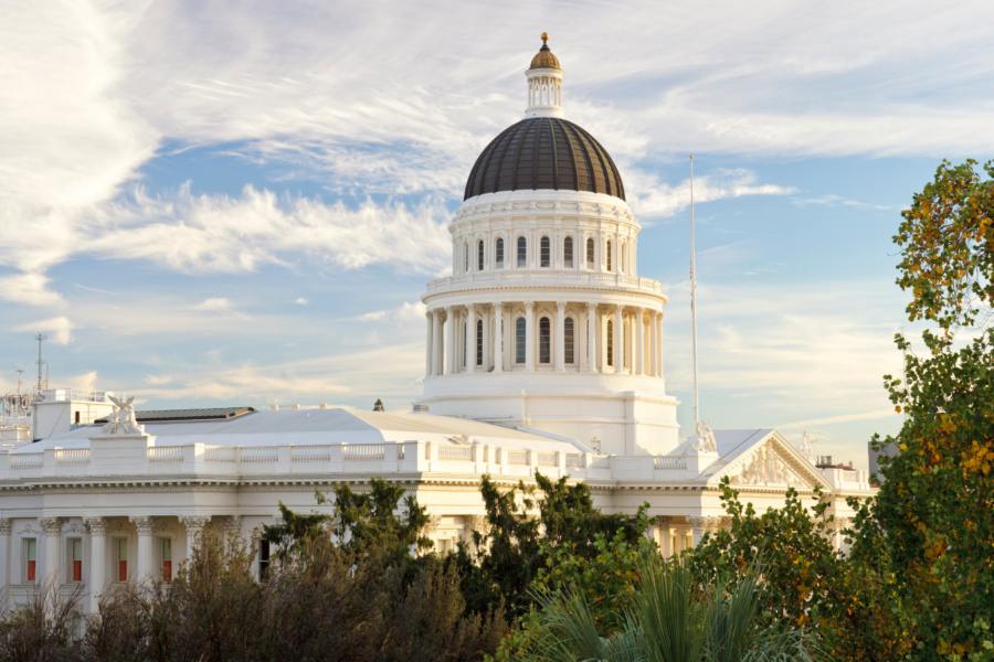 View of the California State Capitol building in Sacramento under a bright sky with wispy clouds, framed by trees and greenery in the foreground.